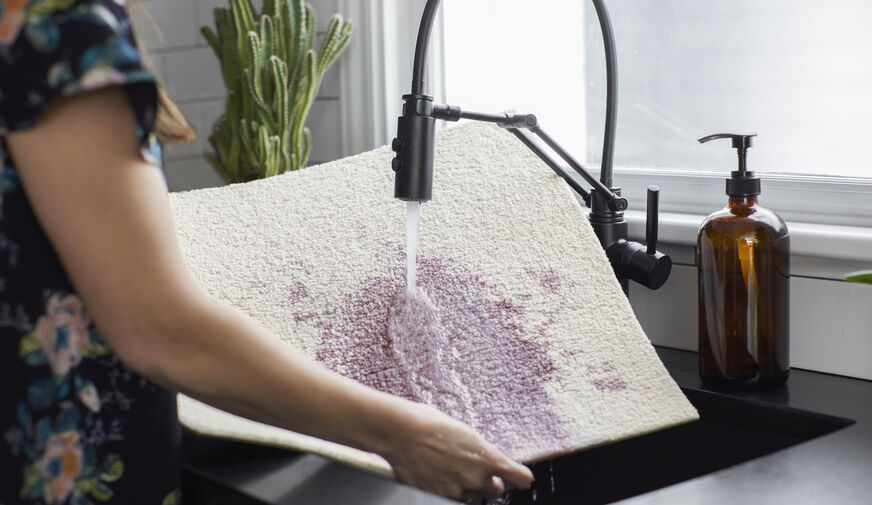 Person in a short-sleeved floral shirt rinsing a white FLOR carpet tile stained with red wine in the sink. 